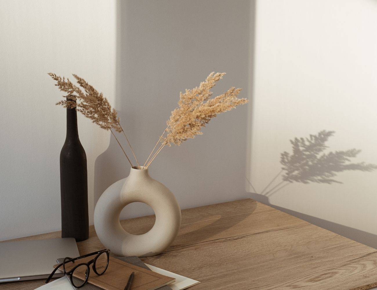 A black skinny vase and a white donut-shaped vase filled with dried plants are sitting atop a wooden desk, alongside a laptop, a pair of glasses, and a notebook.