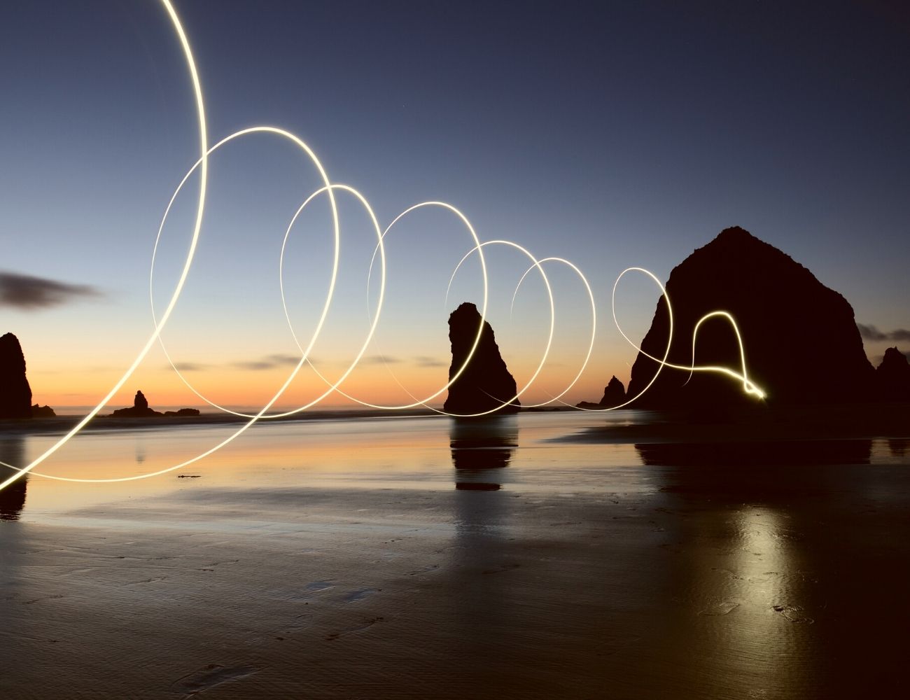 At dusk or dawn, there is a spiral of light from a camera affect in the foreground, in the background there is a beach dotted by large and tall rocks in still water. The sky is a mix of orange, red and blue.