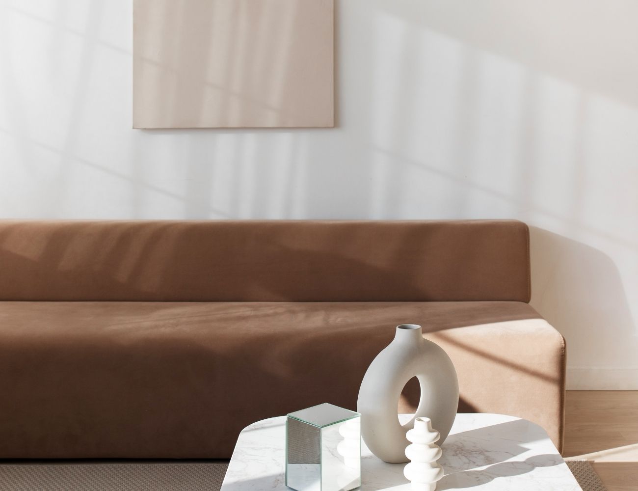 A sleek brown sofa is in the background against a white wall. Two white geometric vases and a glass prism are sitting atop a white marble coffee table in the foreground.